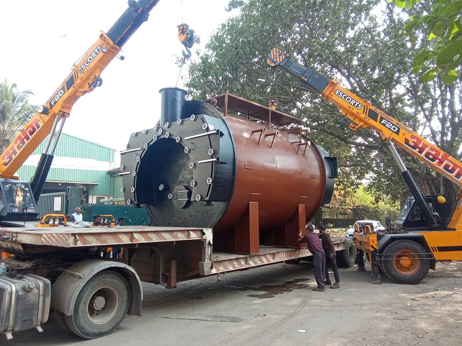 A rust-red industrial boiler lifted by two yellow cranes onto a trailer