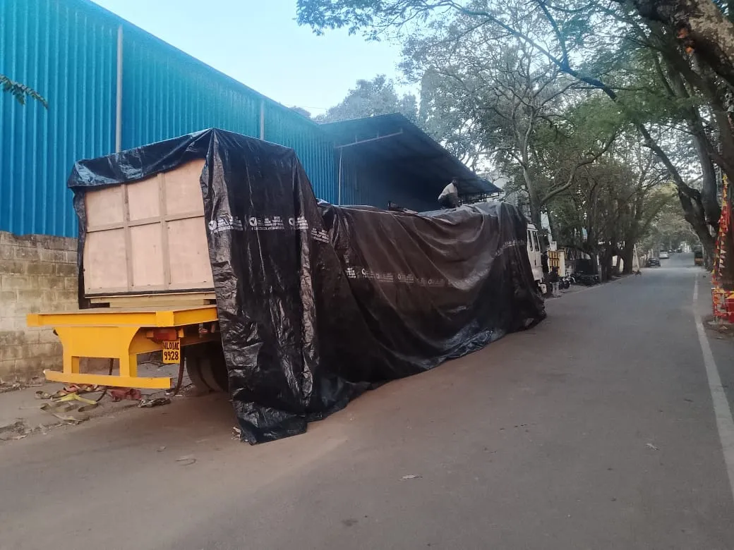 Tarp-covered crated cargo on a yellow trailer, parked on a tree-lined street beside a blue warehouse