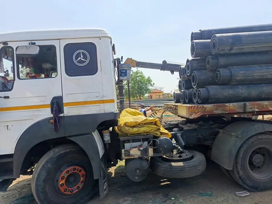 White Mercedes truck cab coupled to a trailer stacked with large black pipes, with a crane loading behind
